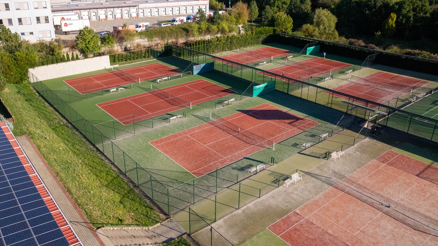 Venkovní kurty areálu Wilson z výšky — několik řad hracích ploch se zeleným kobercem a solárními panely